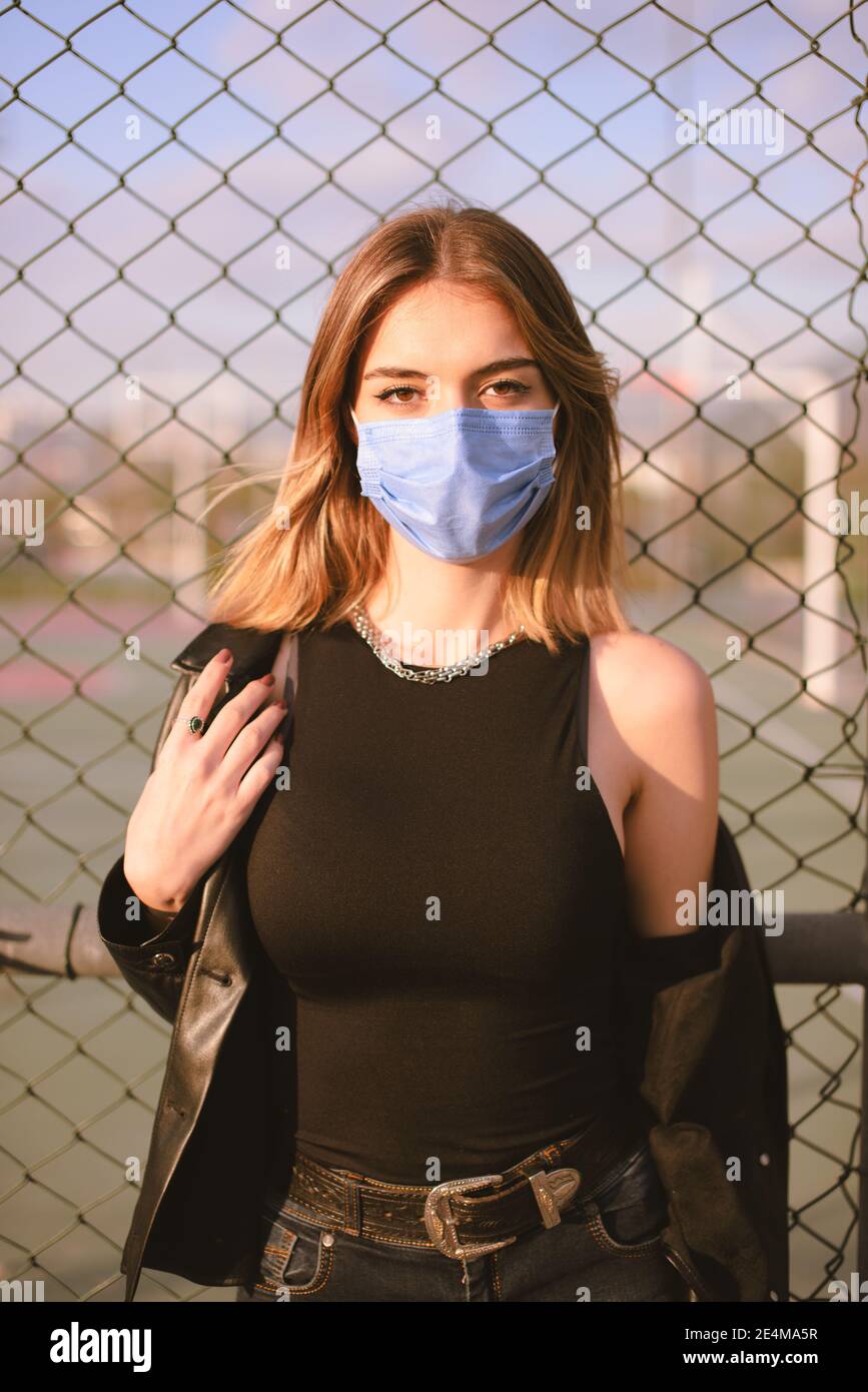 Retrato de una adolescente con una máscara médica en la calle, mirando la cámara y inclinándose hacia una valla. Foto de stock