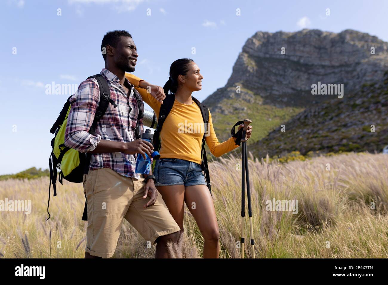 Fit afrcan pareja americana con mochilas marcha nórdica con bastones agua potable en la montaña co Foto de stock