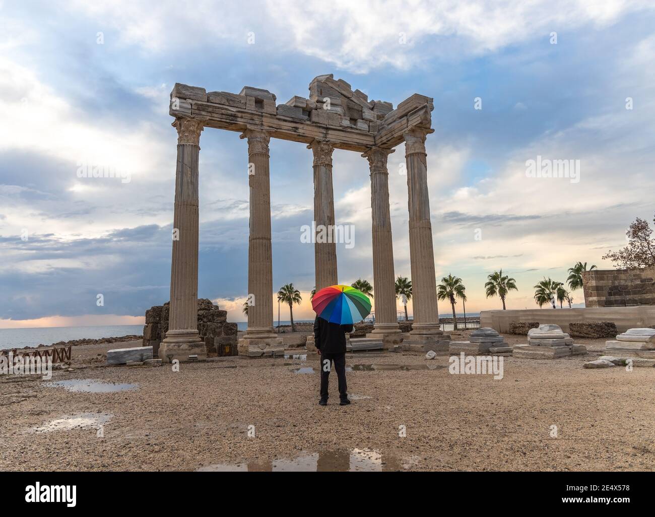 Un hombre vestido de negro con un paraguas de colores arcoiris se encuentra frente al Templo de Apolo en el lado antiguo de la ciudad en el día lluvioso y nublado. Foto de stock