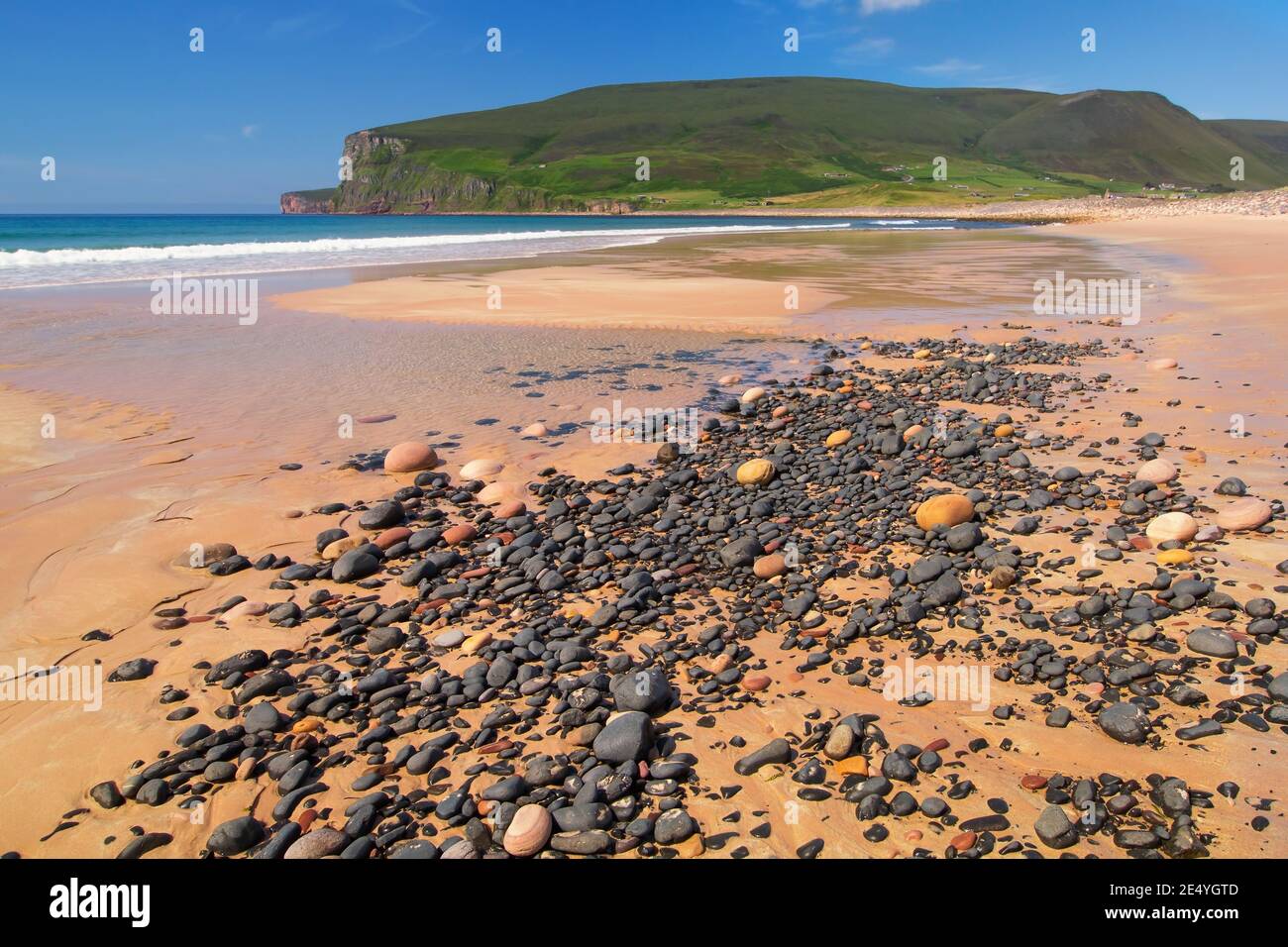 Arena naranja y piedras grises en la playa escocesa con verde colinas y acantilados con cielo azul en la costa de la isla de Orkney Foto de stock
