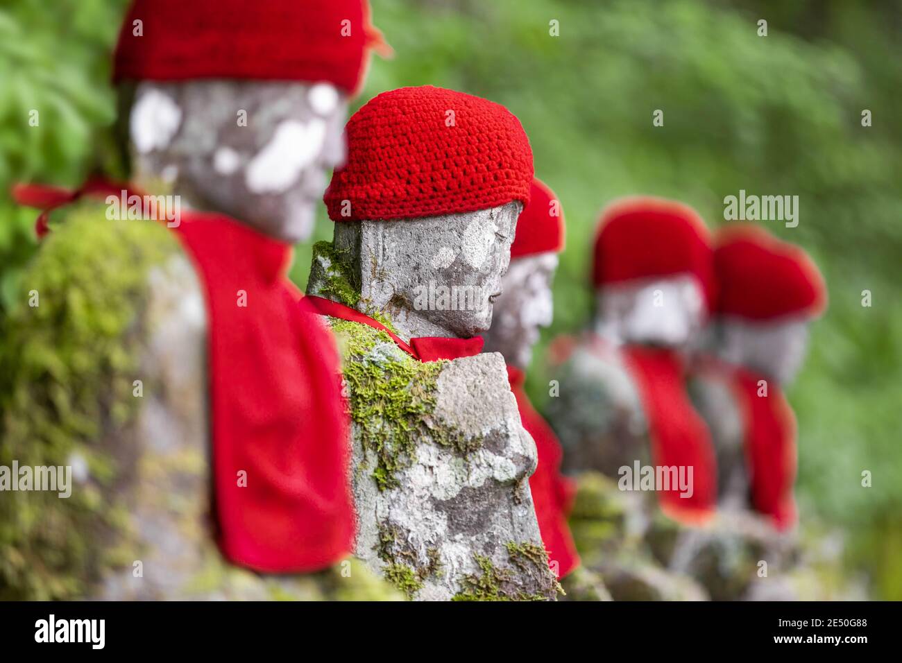 Primer plano de una fila de estatuas de piedra jizo japonés con gorras rojas y baberos, contra un fondo de bokeh verde Foto de stock