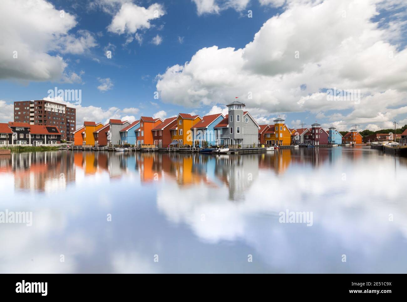 Vista simétrica de un amplio ángulo de un grupo de coloridas casas holandesas modernas frente al agua, bajo un cielo azul de verano con nubes hinchadas Foto de stock