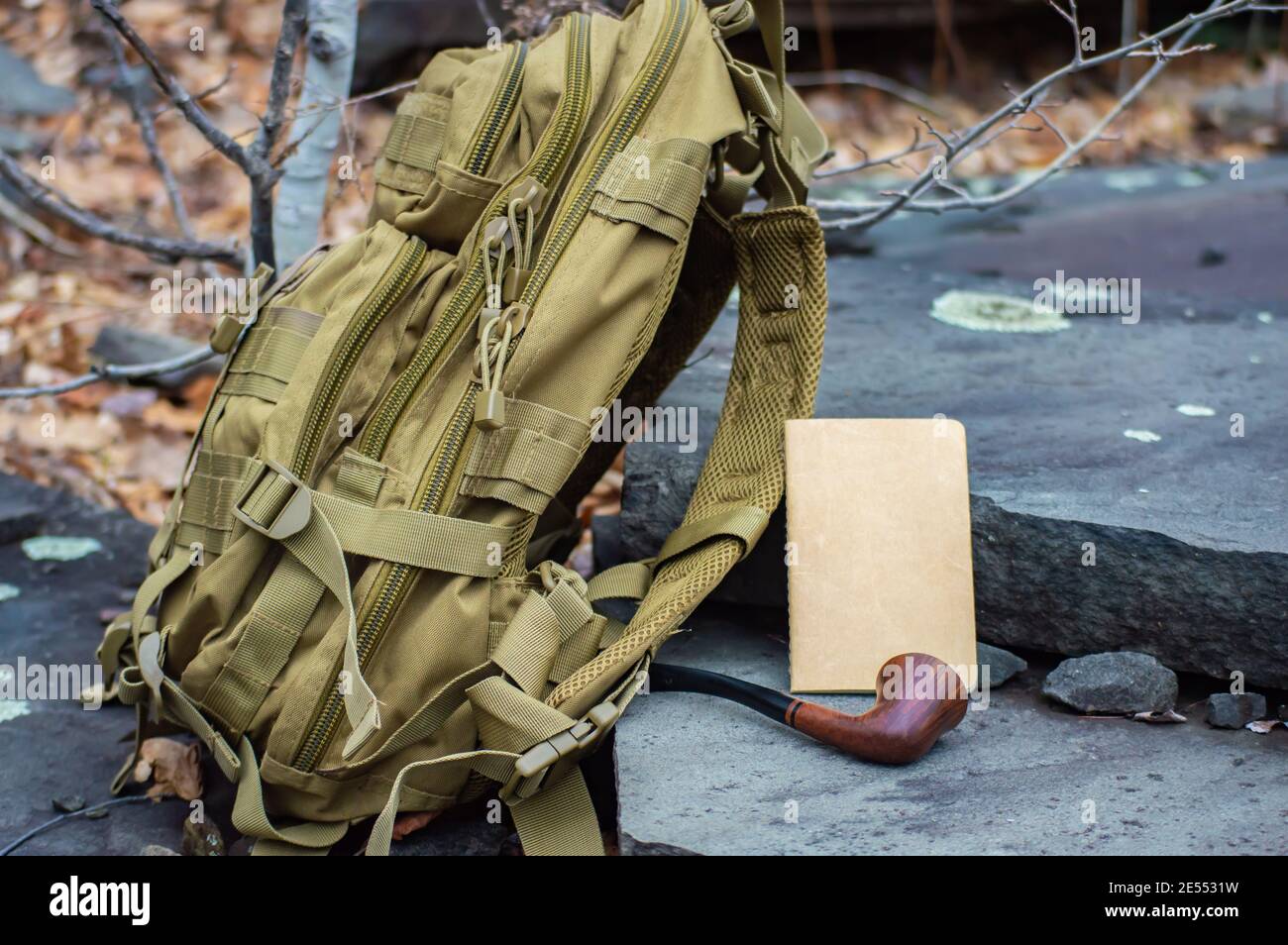 Primer plano de estilo publicitario de una mochila táctica del ejército  verde, un cuaderno de papel y una pipa de época dispuesta en los escalones  de piedra en los bosques Fotografía de