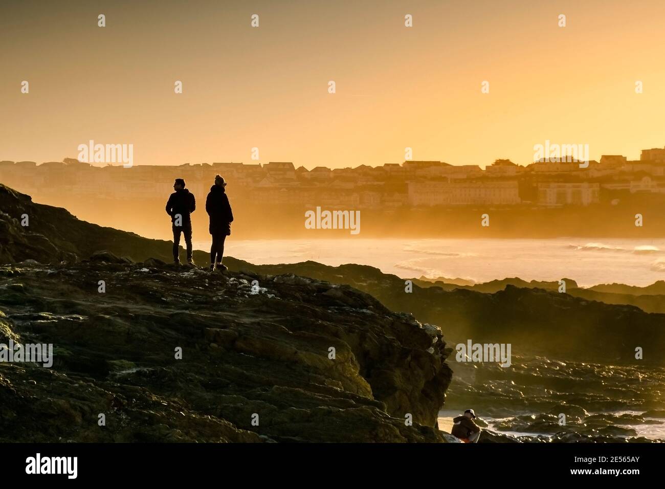 La gente de pie en las rocas se silueta por el sol que pone en Little Fistral en Newquay en Cornwall. Foto de stock