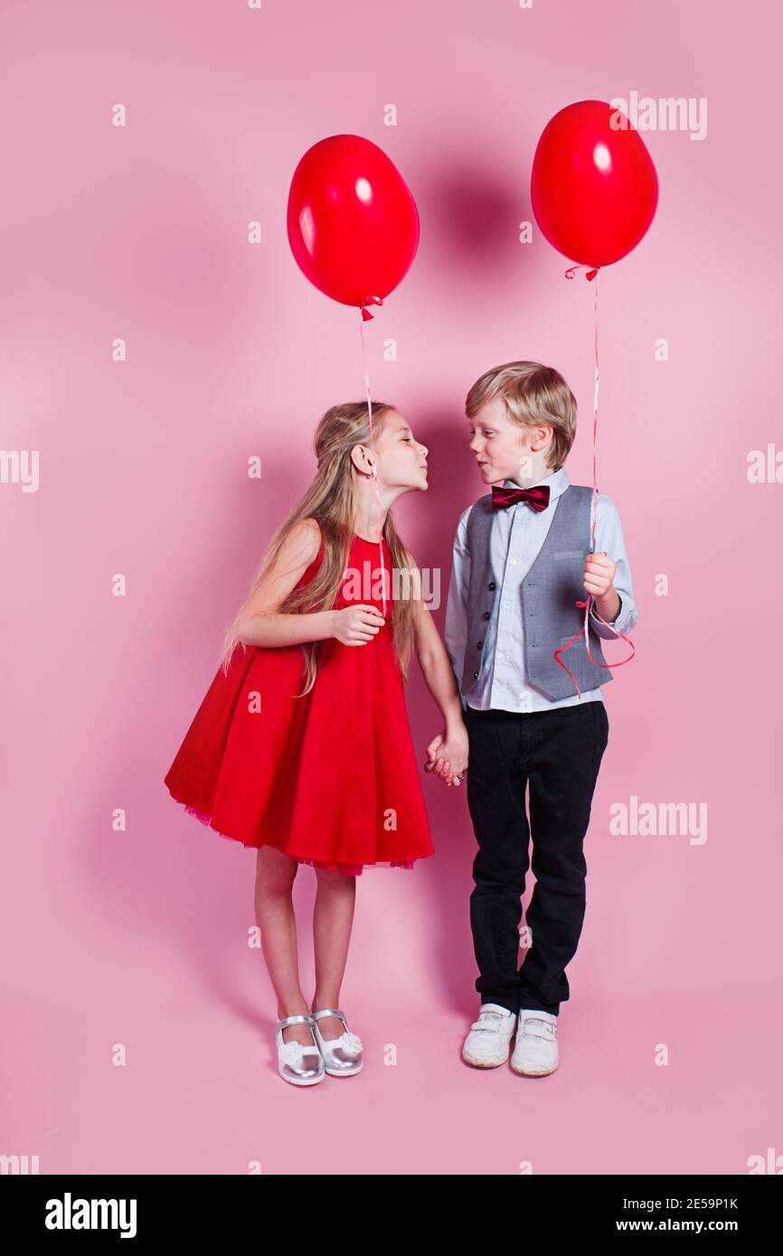 Día de San Valentín. Niño besando a la hermosa niña sobre fondo rosa. Niños sosteniendo globos de corazón Foto de stock