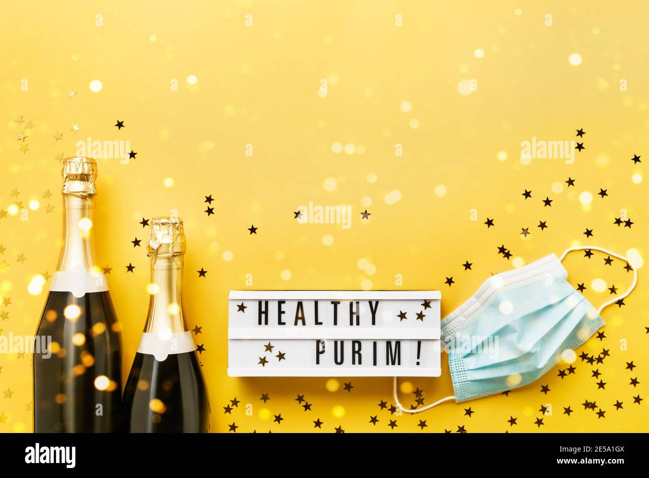 Purim saludable escrito en caja de luz y dos botellas de champán, y máscara médica sobre un fondo amarillo. Lay plano de Purim Carnaval concepto de celebración. Foto de stock