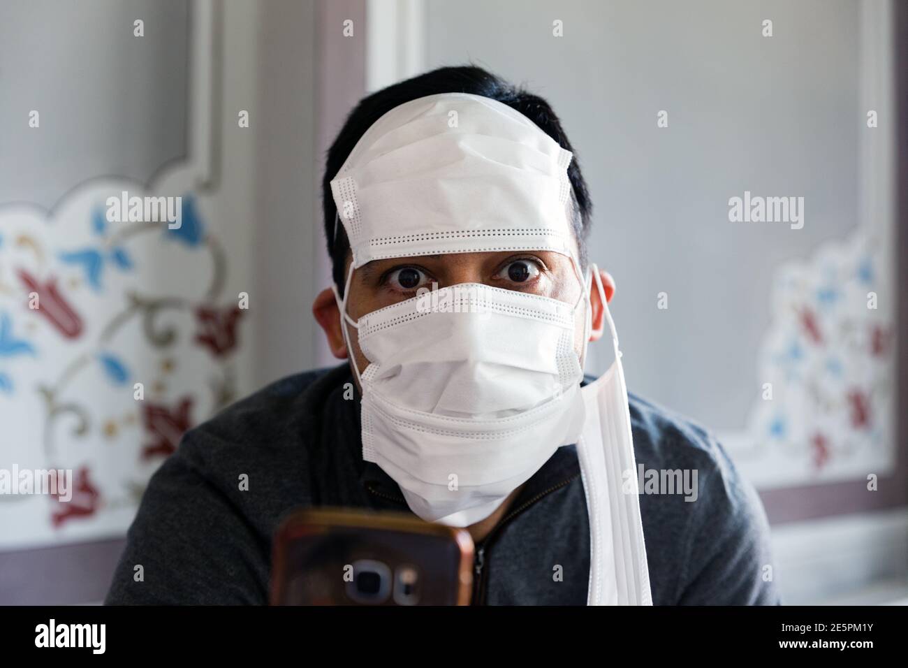 Foto interior retrato frontal de un hombre adulto con pelo corto, mirando la lente con miedo, usando varias máscaras y sosteniendo un smartphone Foto de stock