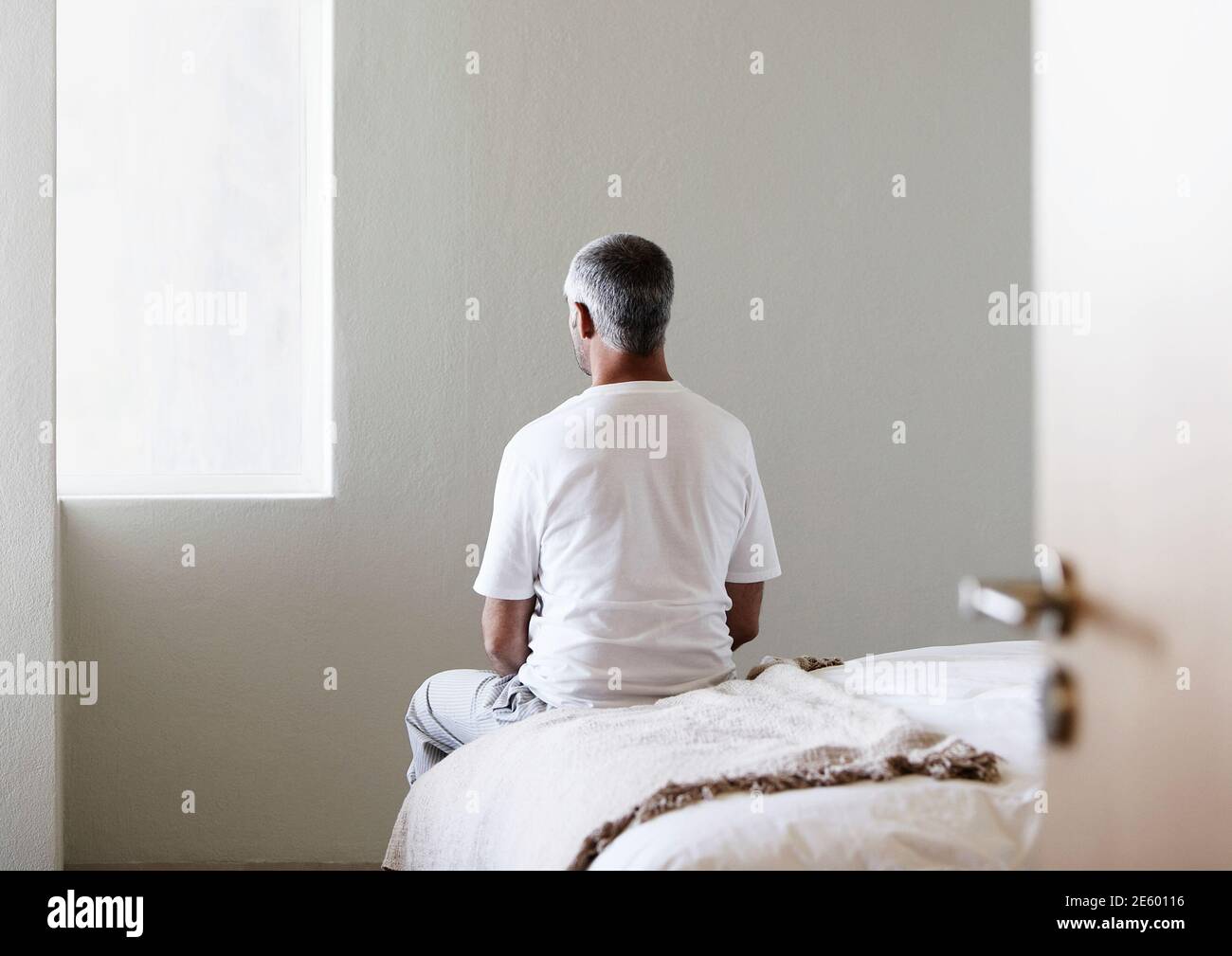 Vista trasera de un varón de mediana edad sentado en el borde de la cama en el dormitorio Foto de stock