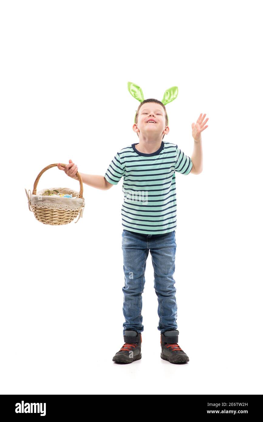Niño joven excitado con orejas de conejo sosteniendo la cesta de Pascua mirando hacia arriba con la mano alzada mugrimacing. Longitud completa aislada sobre fondo blanco. Foto de stock