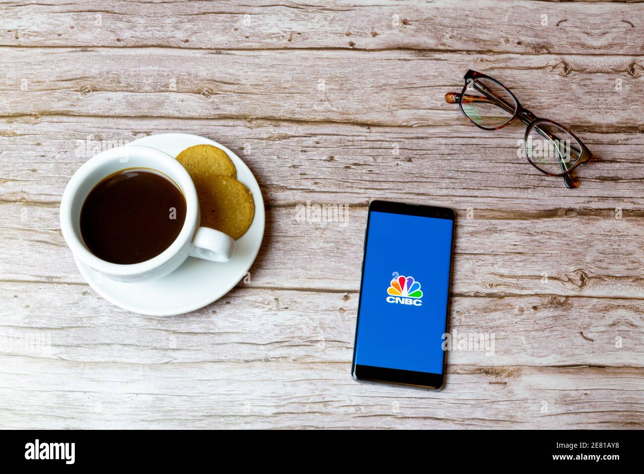 Un teléfono móvil o celular colocado sobre una madera Mesa con una aplicación de noticias CNBC abriendo también un café y gafas Foto de stock
