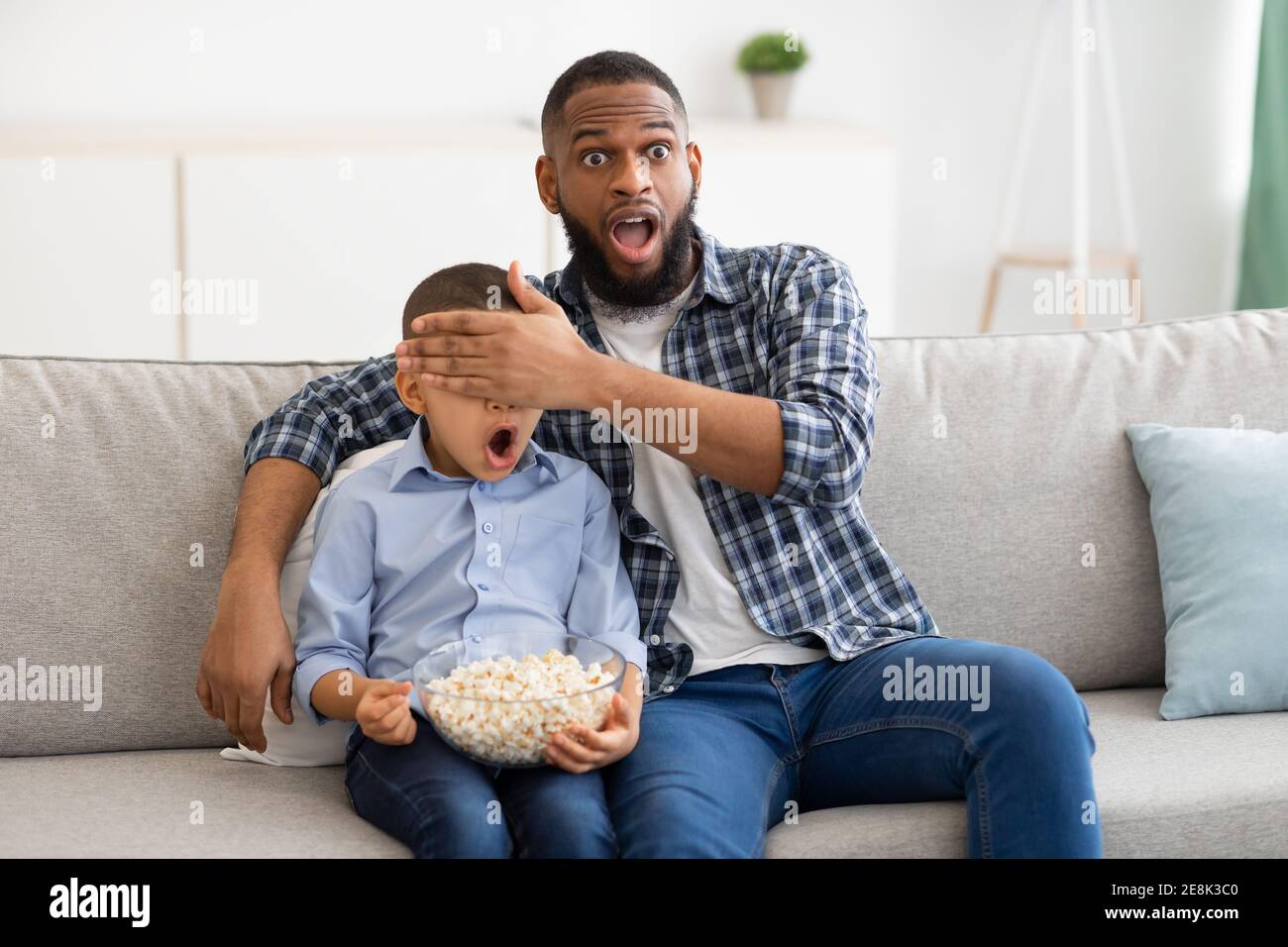 Escandalizado Padre Negro cubriendo los ojos de Hijo viendo la Televisión en el Hogar Foto de stock