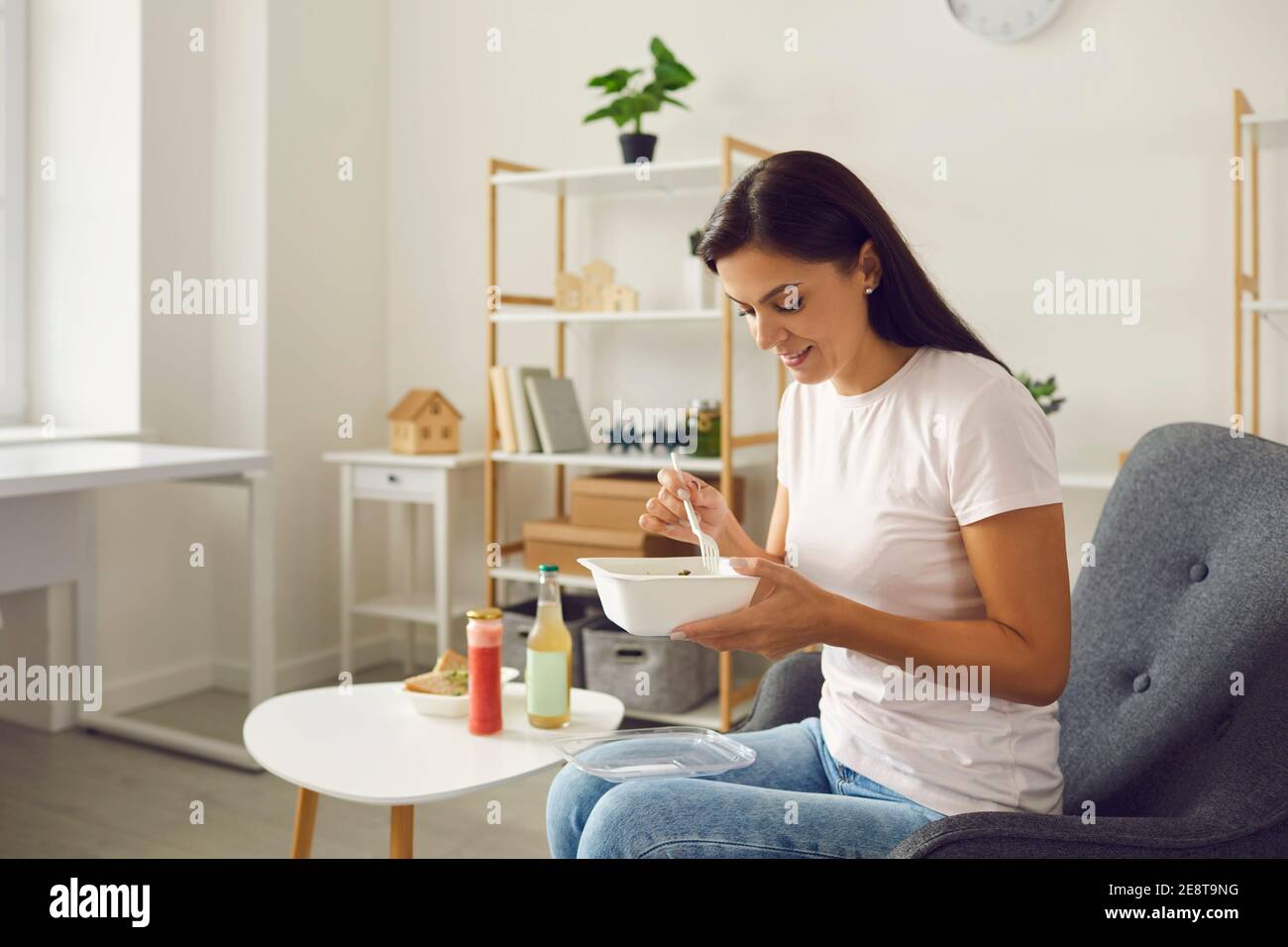 Feliz mujer joven disfrutando de una saludable comida para llevar sentada un sillón en casa Foto de stock