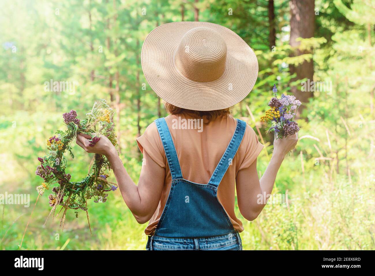 La mujer en un sombrero de paja y un mono vaquero sostiene una corona de  flores del bosque. Vista posterior. Fondo del bosque Fotografía de stock -  Alamy