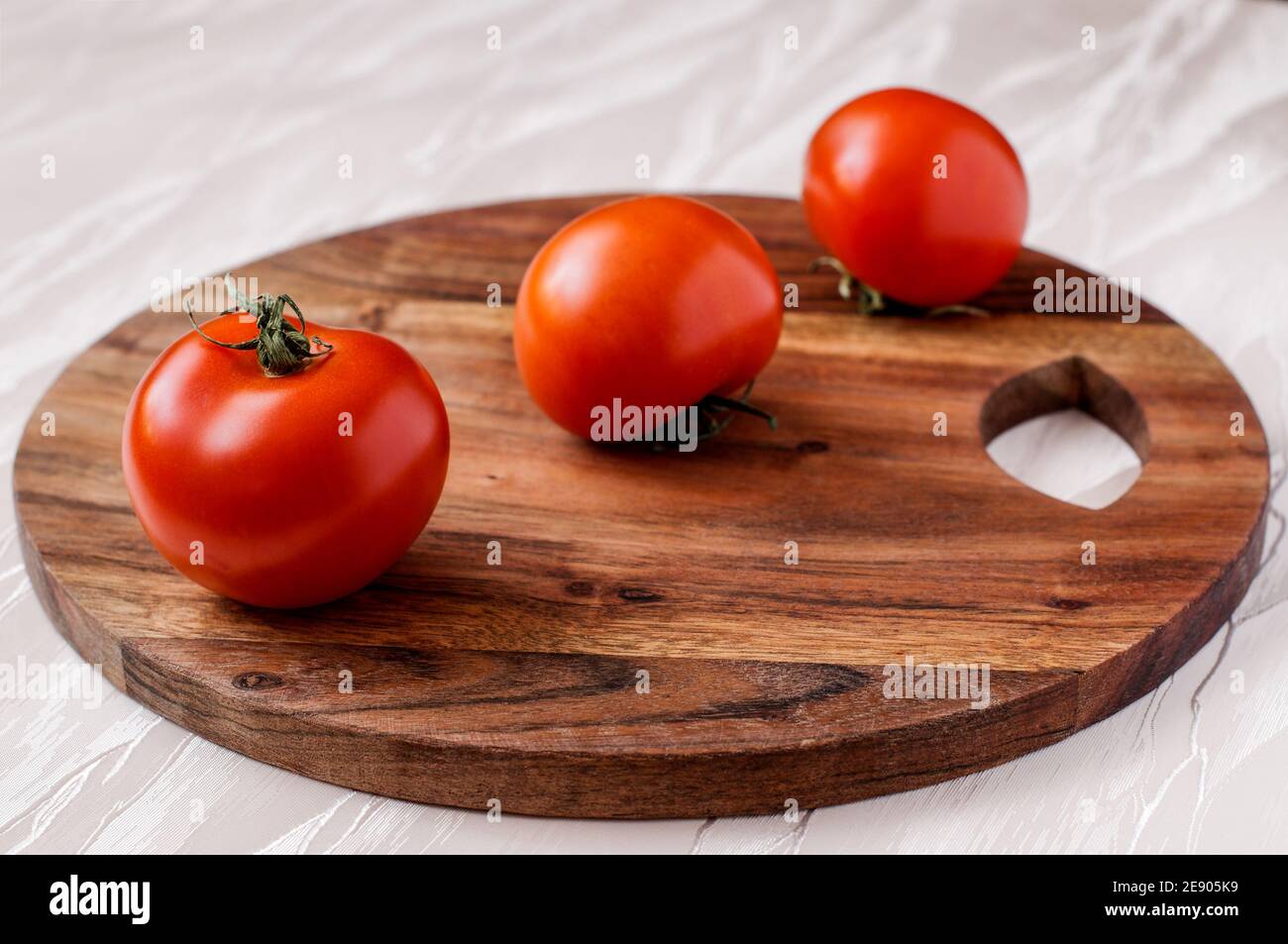 tres tomates rojos en una tabla de cocina redonda de acacia, uno detrás del otro Foto de stock