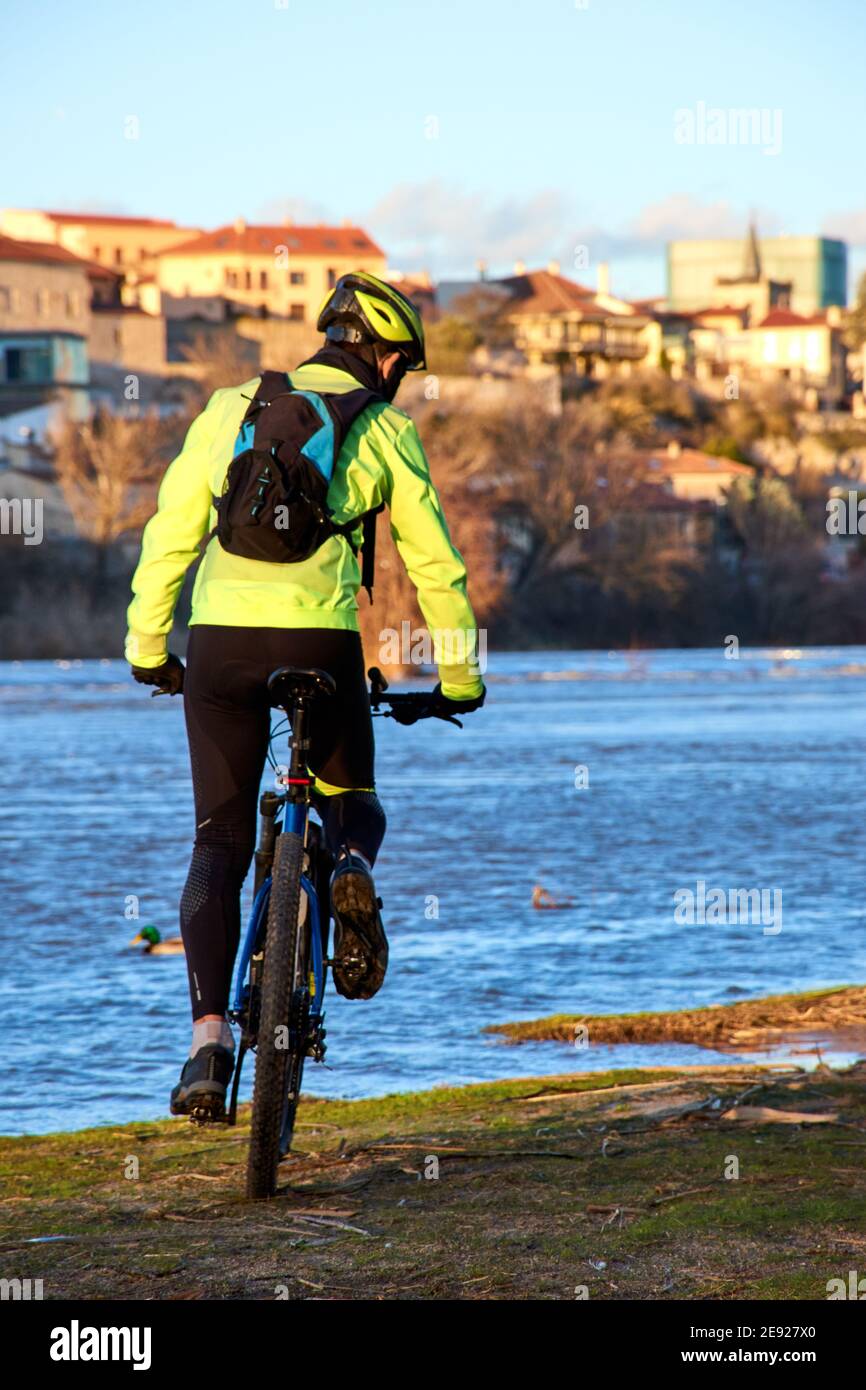 Ciclista de montaña que pasa cerca de una orilla del río en una ciudad.  Vestido con ropa reflectante de colores brillantes, amarillo fluorescente y  negro. MBT al atardecer, e Fotografía de stock -