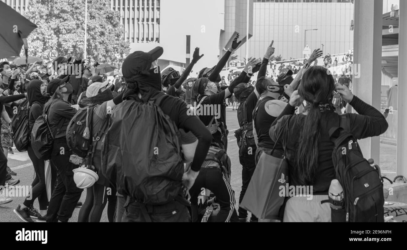 Jóvenes manifestantes en máscaras y sombreros vestidos de negro, se alcanzan en preocupación a medida que dos manifestantes (no visible) subir para eliminar una bandera pro-china. Central, Hong Kong, 1st de octubre de 2019. Foto de stock