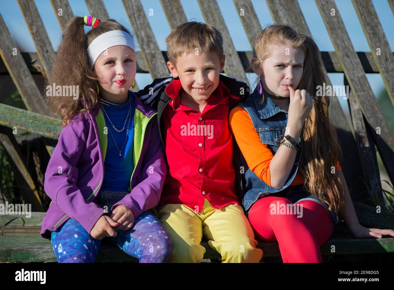 Niños pequeños divertidos: Las chicas con maquillaje brillante vestidas al estilo de los años noventa y un niño con camisa roja están sentados en un banco cerca de una valla sesgada Foto de stock