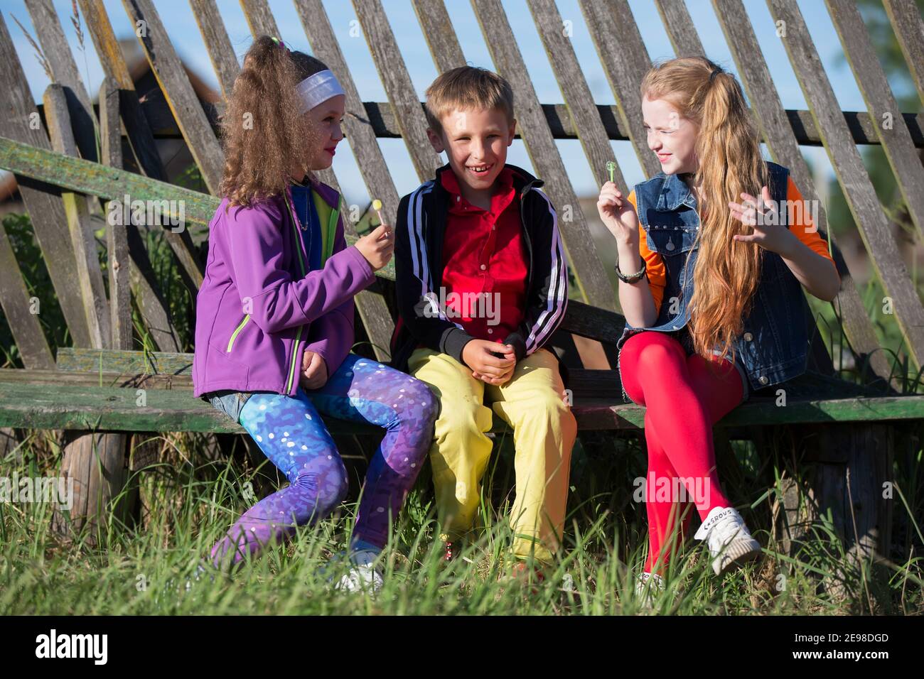 Niños pequeños divertidos: Las chicas con maquillaje brillante vestidas al estilo de los años noventa y un niño con camisa roja están sentados en un banco cerca de una valla sesgada Foto de stock