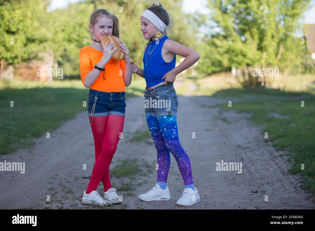 Dos niñas con maquillaje brillante vestido al estilo de los años noventa están comiendo un bollo. Los niños de la aldea rusa. Foto de stock