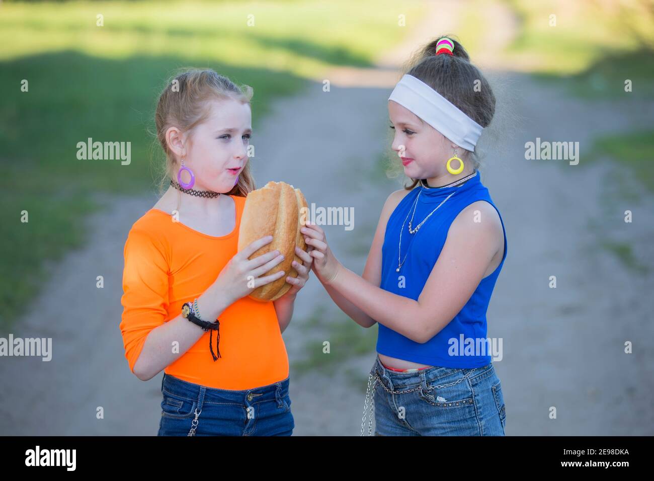Dos niñas con maquillaje brillante vestido al estilo de los años noventa están comiendo un bollo. Los niños de la aldea rusa. Foto de stock
