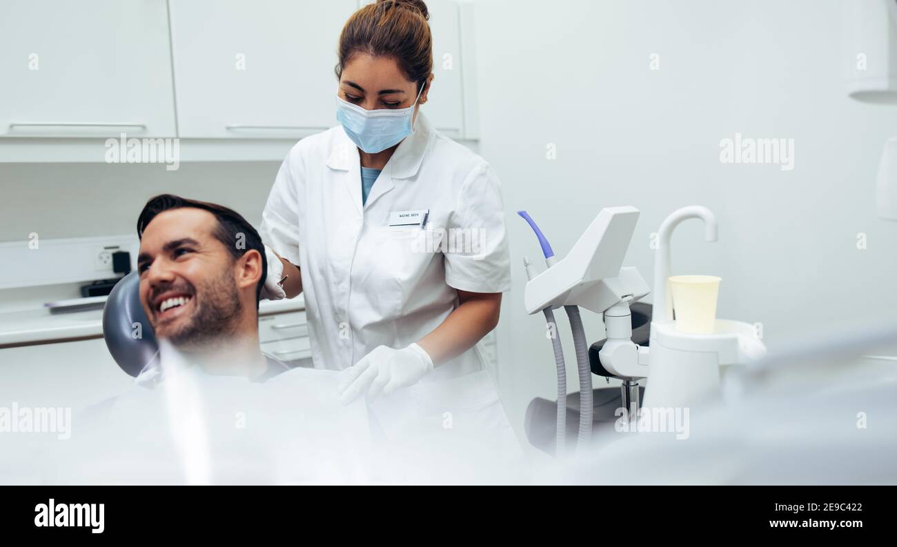 Paciente varón sentado en la silla del dentista mirando hacia fuera y sonriendo con una enfermera que pone en delantal dental sobre el paciente. Dentista que ayuda a un paciente Foto de stock