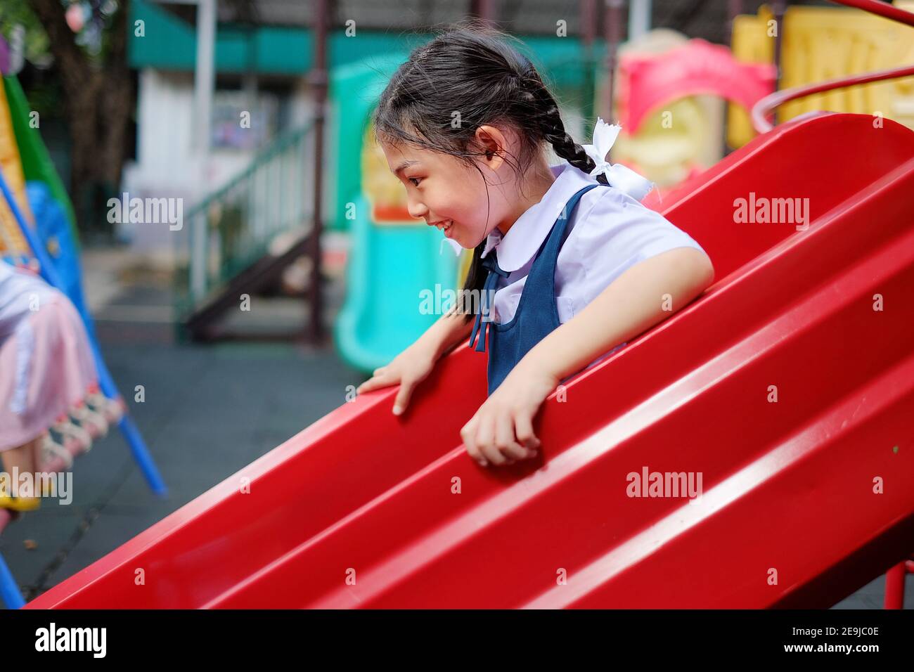 Una linda niña asiática en el uniforme escolar se desliza por un tobogán de plástico rojo en el patio de recreo de la escuela durante el recreo. Foto de stock
