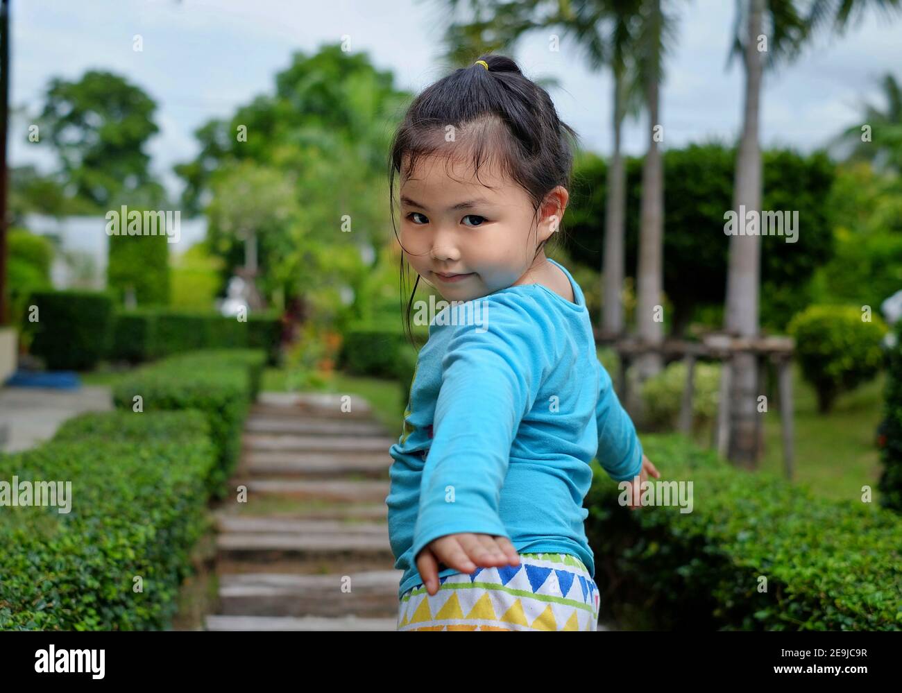Una linda joven asiática con una camisa azul brillante está paseando en un parque con una pasarela de piedra, dando vuelta a una posando para la cámara después de ser Foto de stock