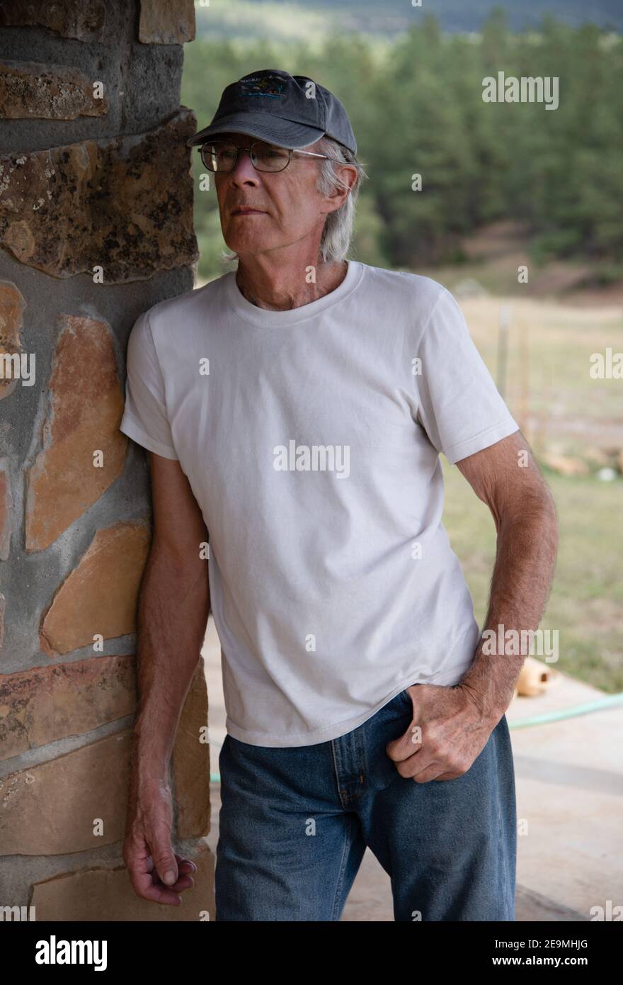 Un joven, hermoso, de 79 años de edad con una camiseta blanca y jeans se apoya contra una pared de piedra. Foto de stock