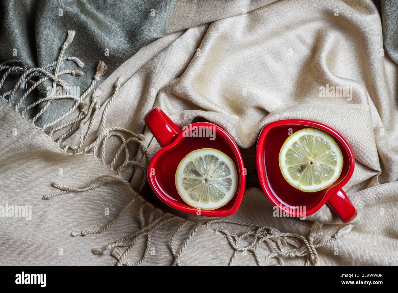 Tazas de té y bufanda en forma de corazón rojo, símbolo del día de San Valentín. Dos bebidas calientes y platos en una acogedora casa. Foto de stock
