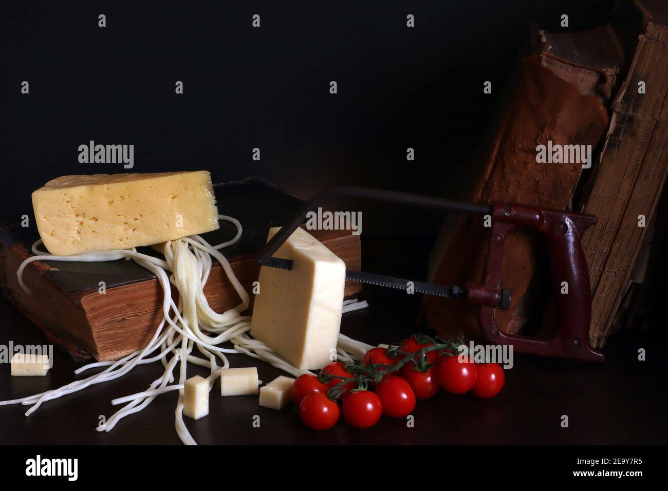 Todavía la vida con tres quesos, tomates cherry, libros viejos y sierra de brazo, foto comida absurda Foto de stock