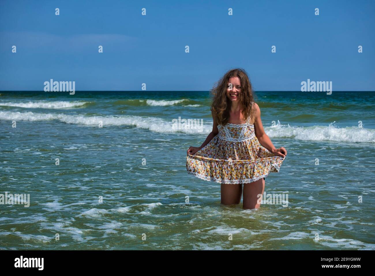 La niña en un vestido floral está de pie en el mar sosteniendo su vestido y riendo Foto de stock