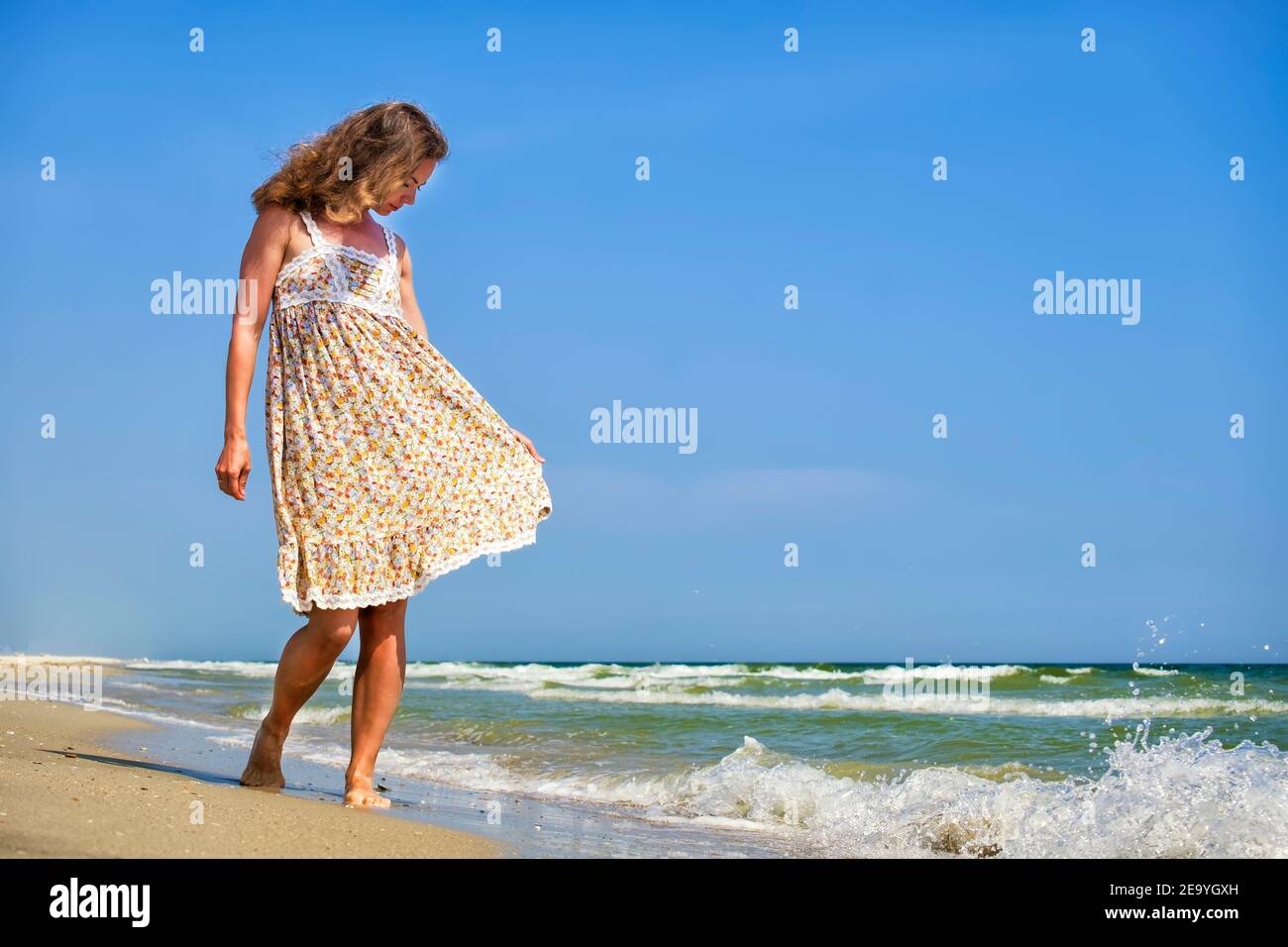 Niña en un vestido floral se pone de pie en la orilla del mar con la cabeza hacia abajo Foto de stock