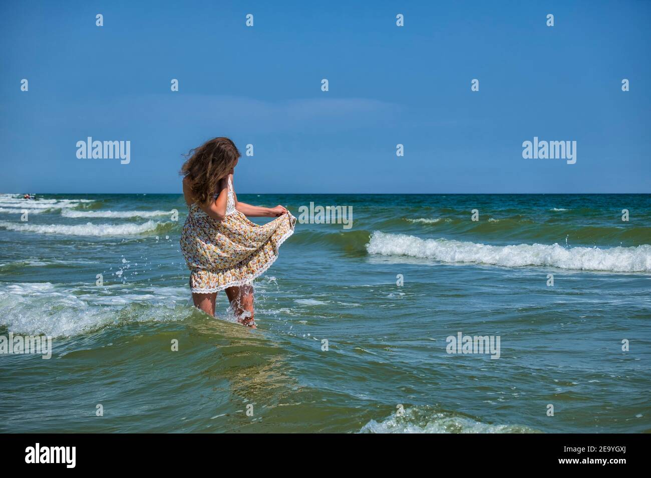 La niña se encuentra en el mar, levantando el borde de una sundress para que no esté húmeda Foto de stock