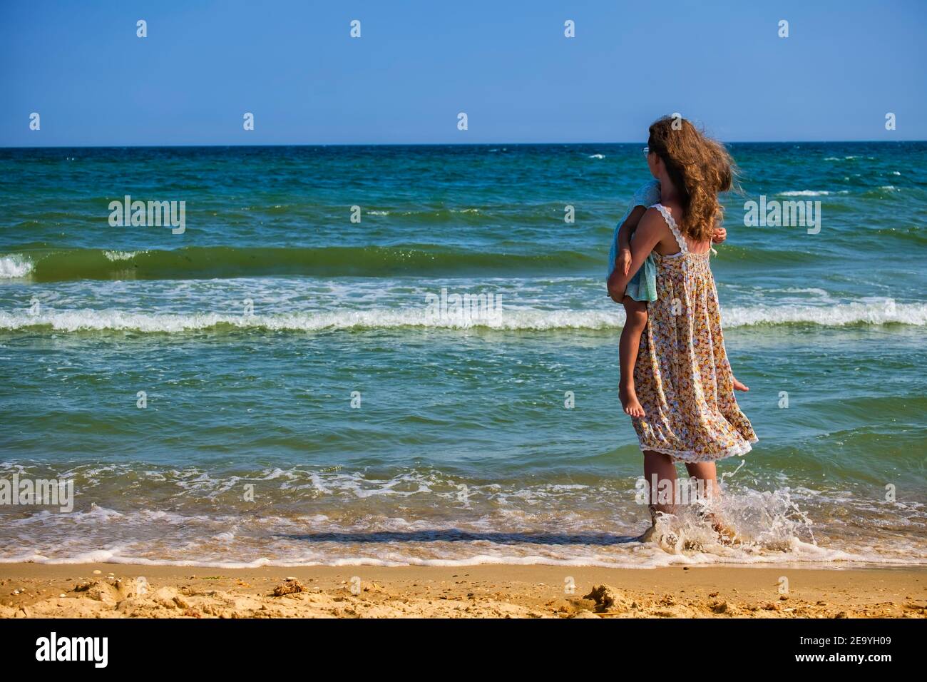 Una niña con un bebé en los brazos se pone de pie la orilla del mar y mira a la distancia Foto de stock