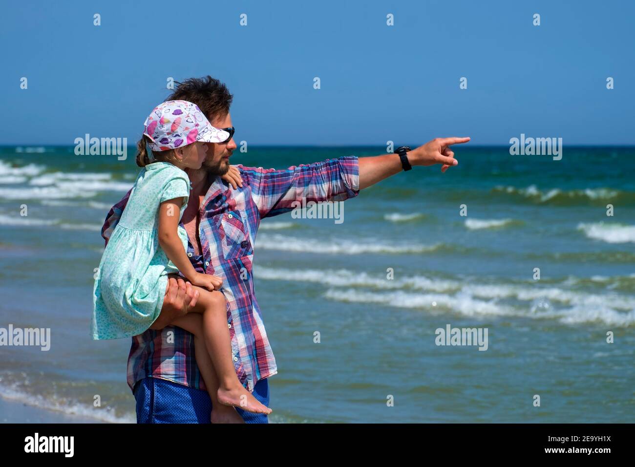 Papá sostiene a su hija en sus brazos y muestra algo muy lejos del mar Foto de stock