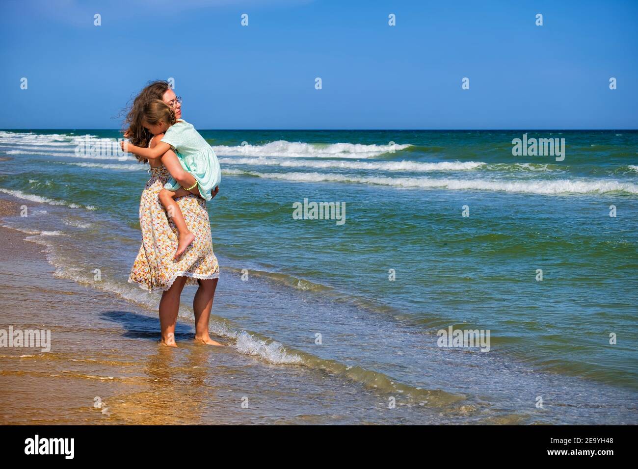 Mamá sostiene a su hija pequeña en sus brazos con amor. Familia joven feliz caminando a lo largo de la orilla del mar Foto de stock