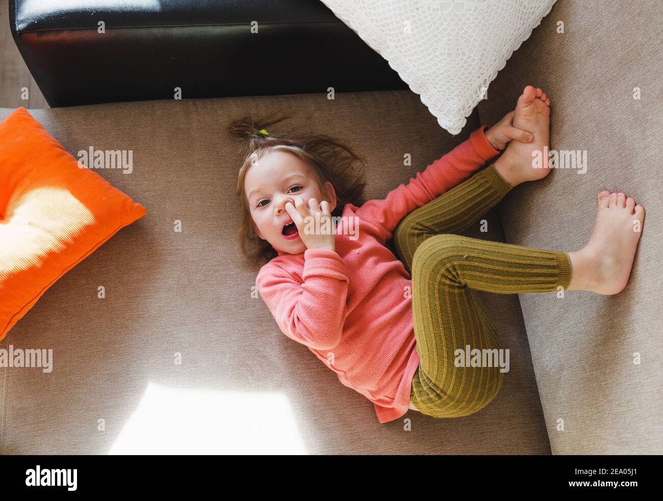 una niña en una sudadera rosa está acostada en el sofá entre las almohadas  con las piernas levantadas y recogiendo la nariz. Juegos en casa Fotografía  de stock - Alamy