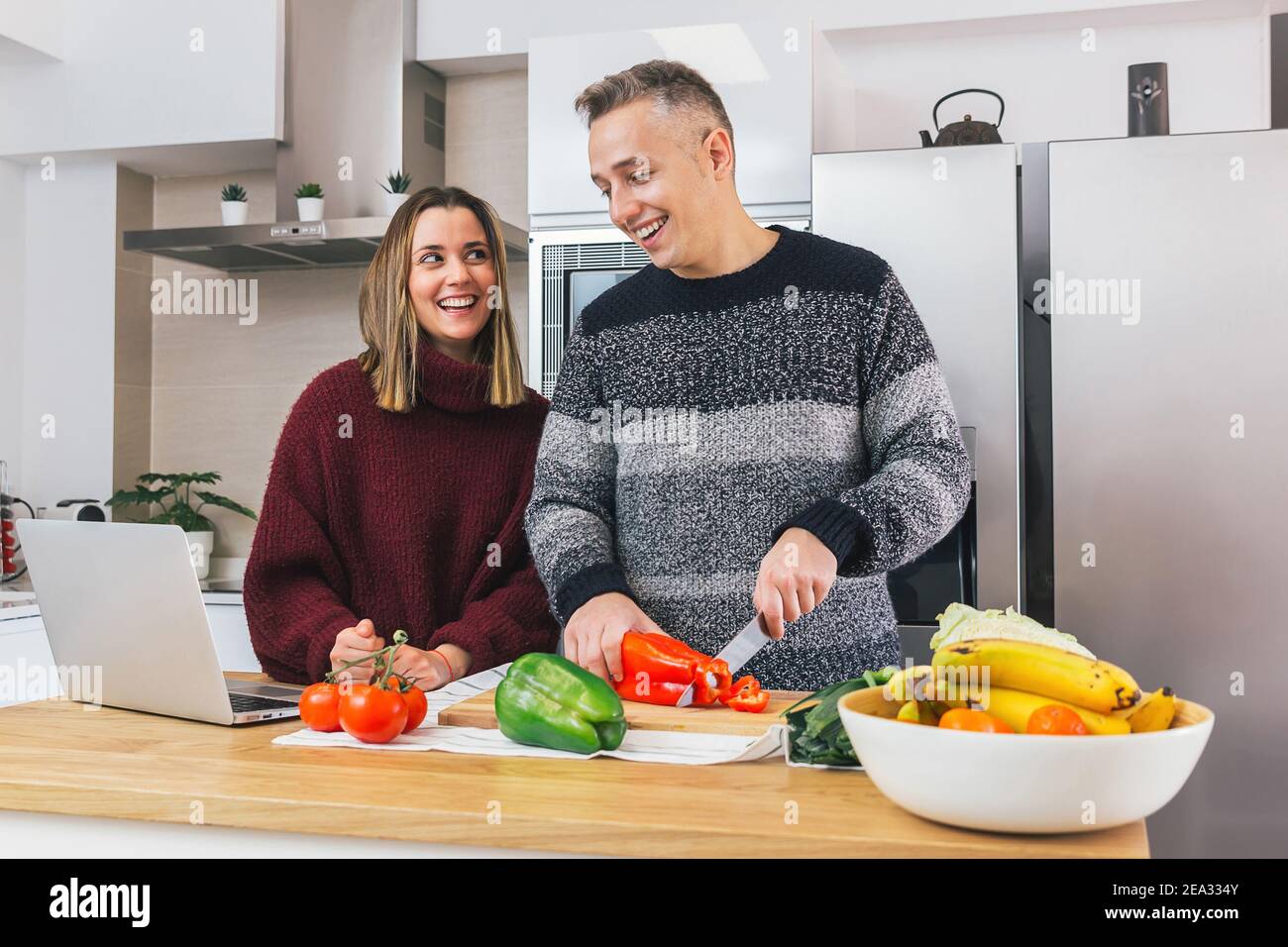 Foto de stock de una joven pareja feliz riendo y preparando comida saludable en su cocina y leyendo recetas en el cuaderno. Aprender a cocinar y disfrutar Foto de stock