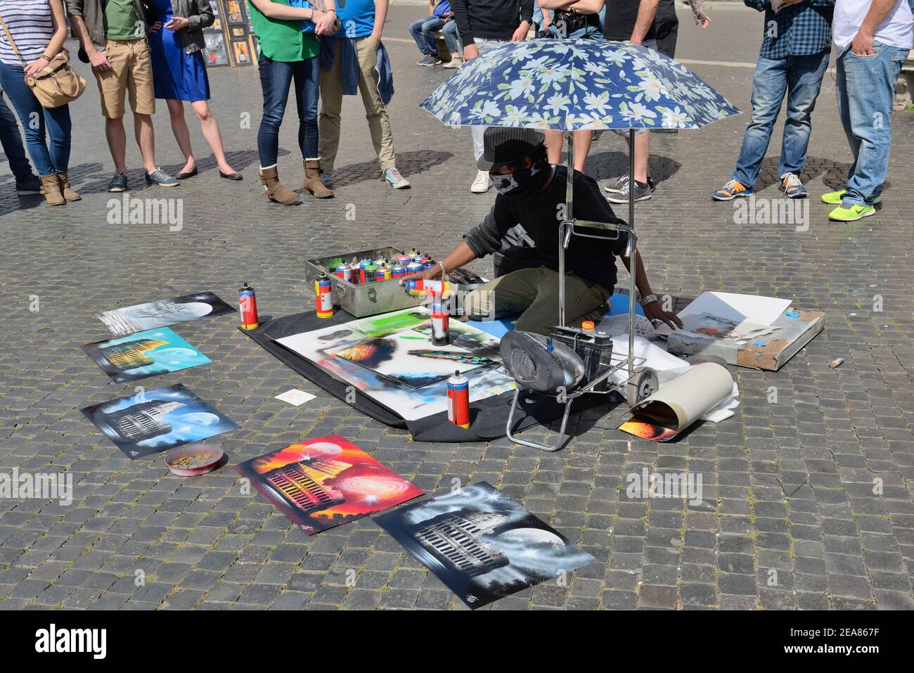 Un hombre joven bajo un paraguas pintando cuadros coloridos usando latas de aerosol de pintura en una calle adoquinada con el turista mirando en Roma, Italia Foto de stock