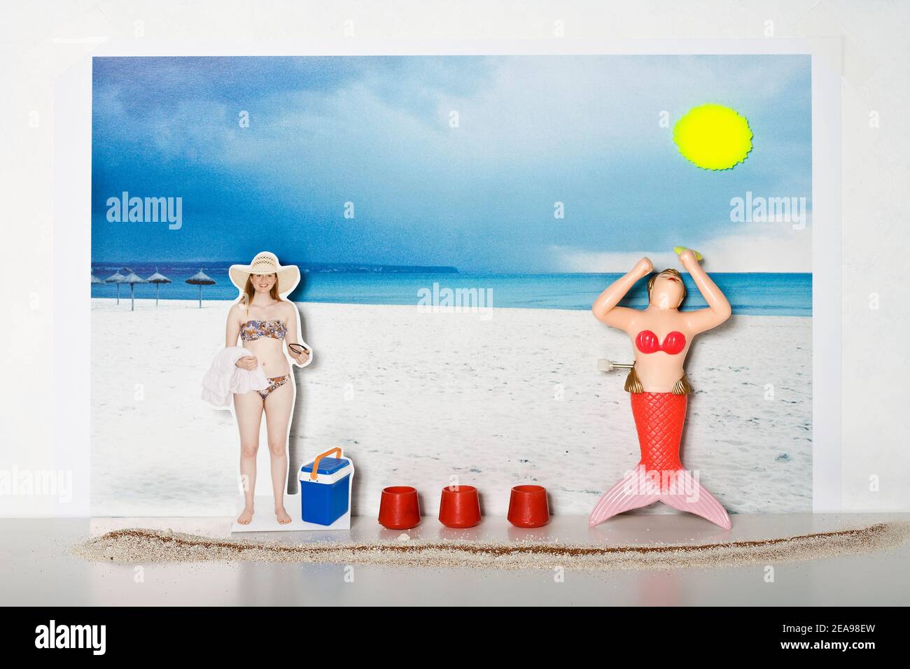 Mujer joven en el bikini se encuentra en la playa, toalla de baño y gafas de sol en la mano, baño de plástico sirena adoran el sol, nevera y contenedor rojo entre ellos, sol de plástico, foto de fondo de una playa, arena subterránea y blanco, foto collage Foto de stock