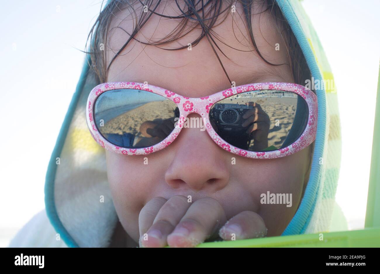 niño de 3 años tomando el sol después de nadar. Está usando una toalla de poncho encapuchada y gafas de sol Foto de stock