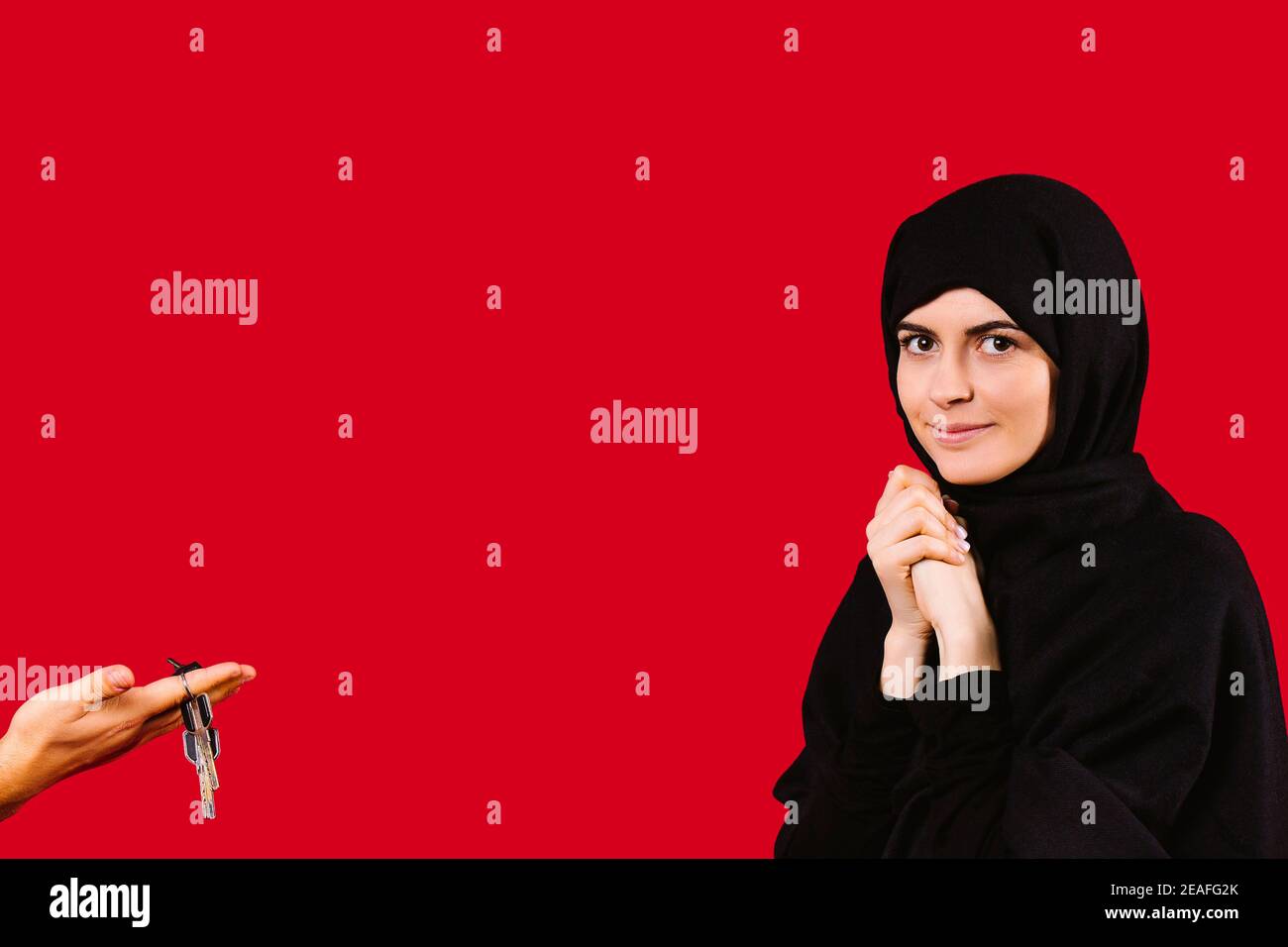 Chica joven sorprendida en negro hijab mira la cámara. La mano de un hombre sostiene las llaves del apartamento. Regalo para mujeres musulmanas sobre fondo rojo. Foto de stock