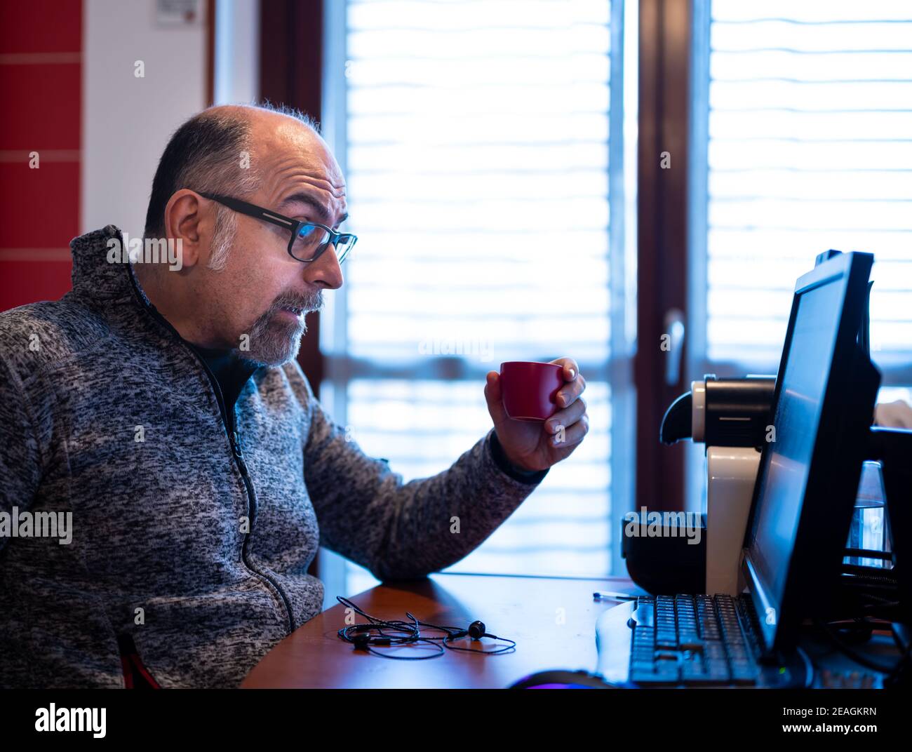 Un hombre caucásico de mediana edad que trabaja desde casa, mientras bebe un café, está agradablemente sorprendido por una notificación que lee en el monitor de su computadora. Foto de stock