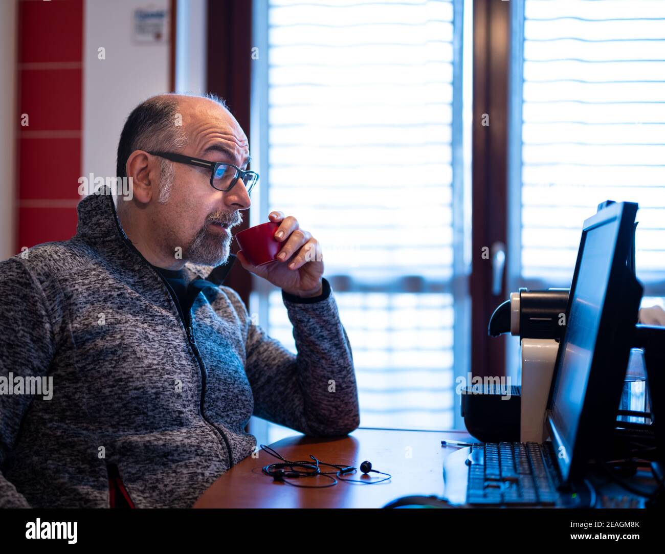 Un hombre caucásico de mediana edad que trabaja desde casa, mientras bebe un café, está agradablemente sorprendido por una notificación que lee en el monitor de su computadora. Foto de stock