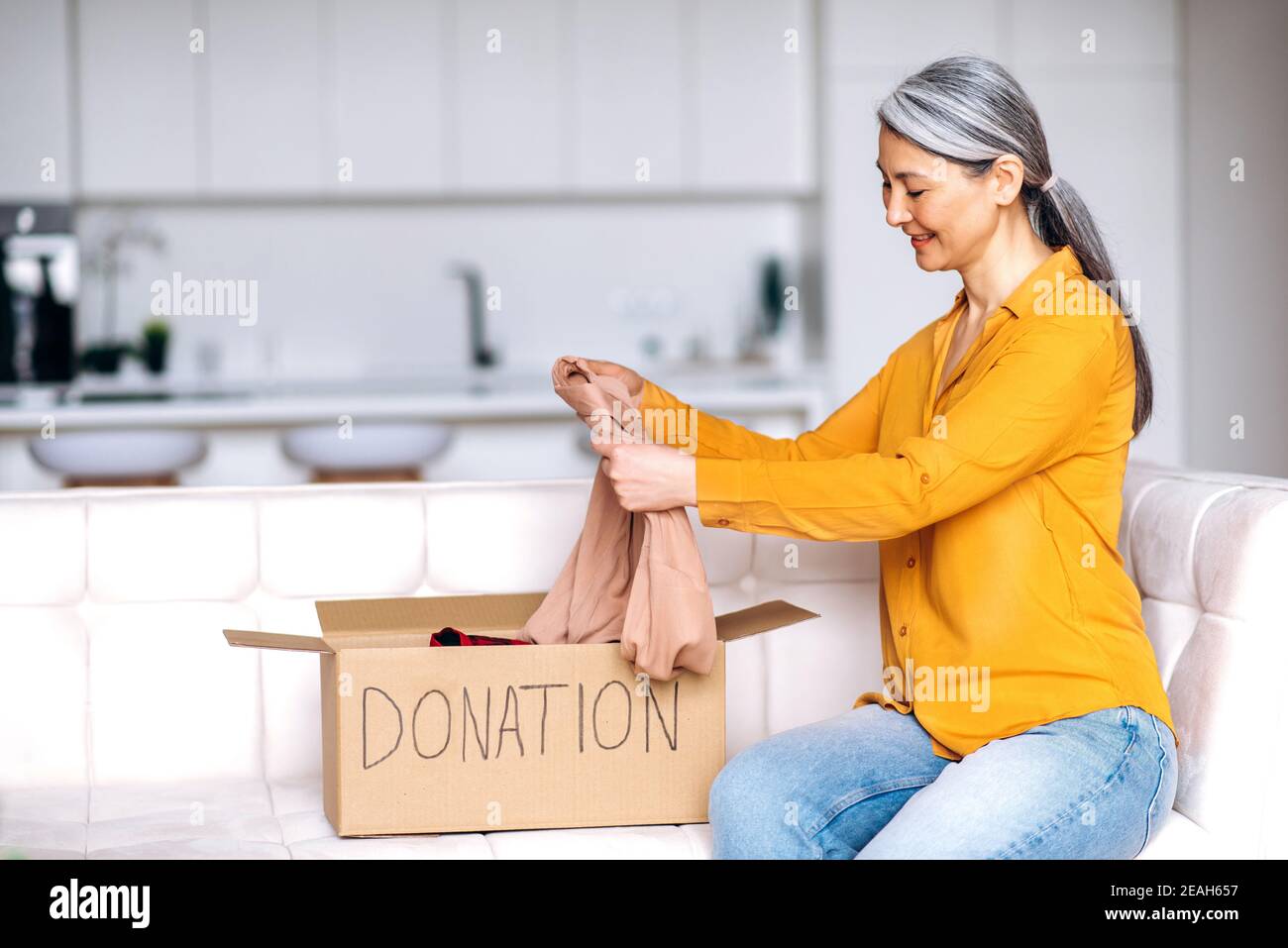 Caridad, donación. Feliz mujer mayor de pelo gris sentada en casa en el  sofá desempaqueta la caja de donación con de ropa, para su reutilización.  Ayudar a las personas pobres y necesitadas