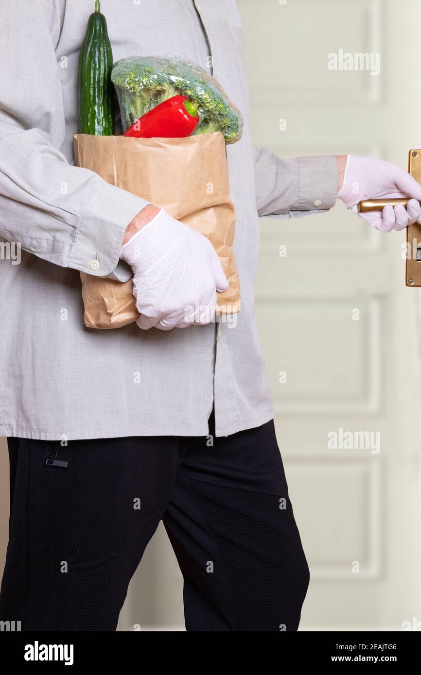 Un hombre que lleva guantes médicos y una bolsa de compras con verduras frescas entra en la casa. Concepto de fortalecimiento del sistema inmunitario y protección contra las infecciones por coronavirus. Foto de stock