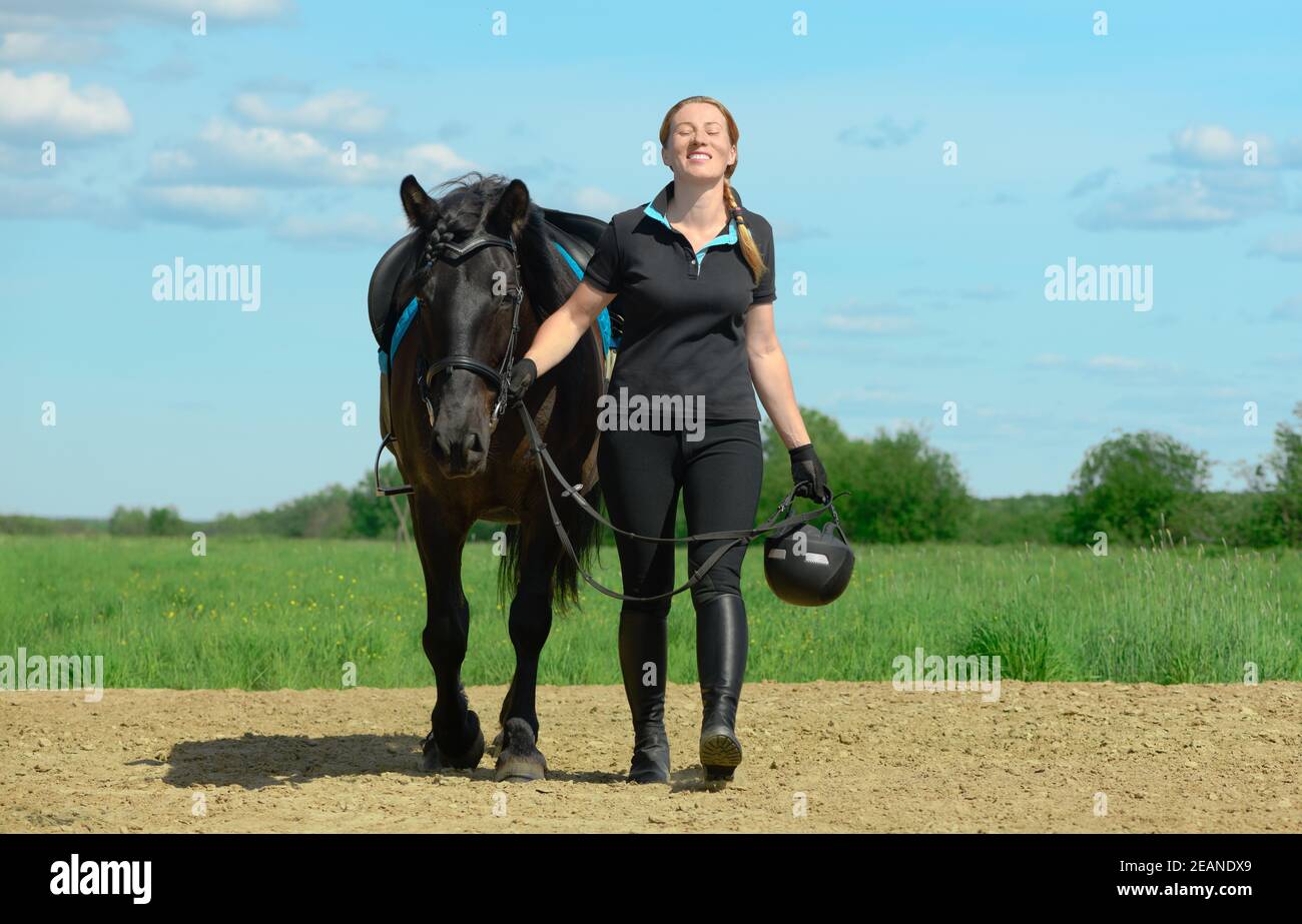 Una mujer caucásica sonriente, de 40 años de edad, sostiene las riendas de su caballo y camina por el suelo arenoso al aire libre. Foto de stock