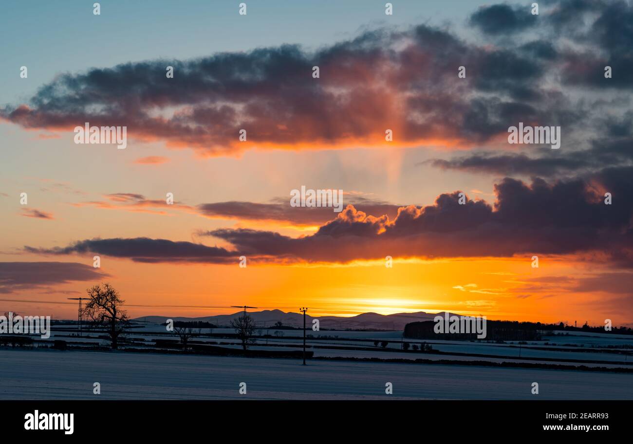 East Lothian, Escocia, Reino Unido, 10 de febrero de 2021. Reino Unido Clima: Un cielo naranja ardiente al atardecer sobre un paisaje cubierto de nieve mirando hacia el contorno de las colinas Pentland en la distancia Foto de stock