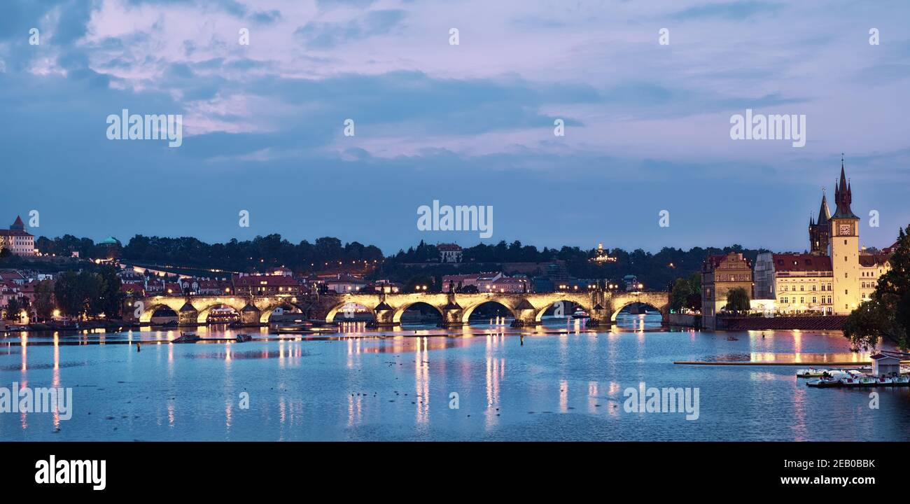 Río Praga al atardecer al atardecer. Imagen panorámica del Puente Carlos iluminado y edificios junto al río con una torre de reloj Foto de stock