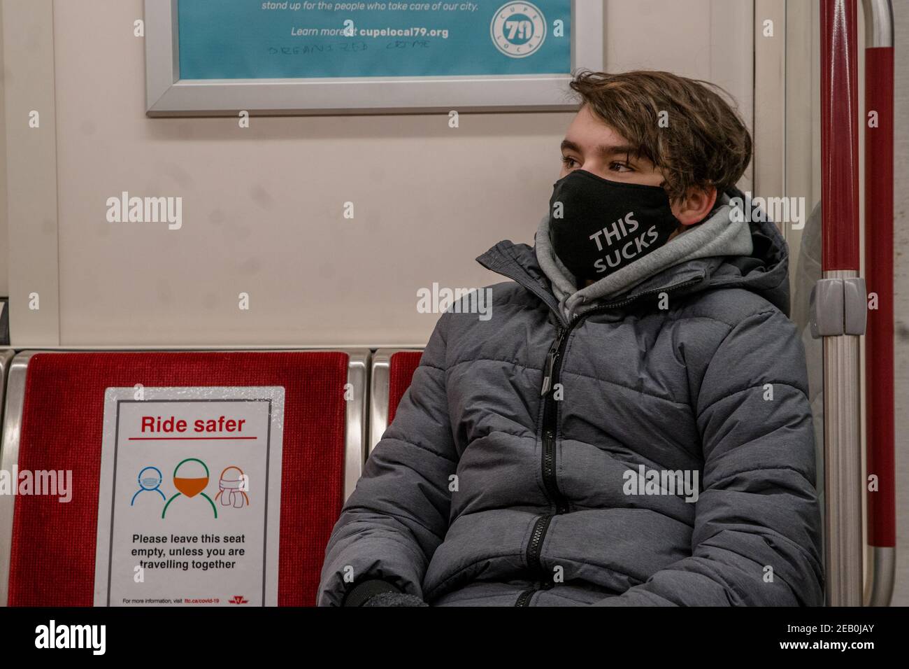 El adolescente lleva una máscara que dice "ESTO CHUPA" en el metro durante  la pandemia de COVID-19. Señalizaciones en el asiento al lado de él tiene  instrucciones para distanciarse social Fotografía de